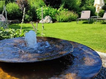 Rundes dunkles Wasserbecken mit sprudelndem Quellstein vor einer Rasenfläche mit Stauden und Gartenliegen