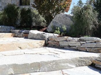 Massive Natursteinstufen mit Trockenmauer, großen Findlingen und Ziergräsern an einem Wohnhaus