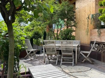 Gemütlicher Gartensitzplatz mit Holztisch und Stühlen, umgeben von Bäumen, Gehölzen und üppigem Grün